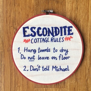 Op de afbeelding: Geborduurde cottage regels in een houten hoepel. De tekst luidt "ESCONDITE COTTAGE RULES" in blauw garen, met extra regels eronder. De hoepel heeft een rode rand en staat tegen een houten achtergrond.