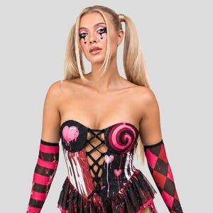 May include: A strapless corset top featuring a black lace-up front, pink heart and swirl graphics, and a ruffled hem. The outfit includes matching arm sleeves with pink and black stripes and diamond patterns, paired with matching leg chaps.