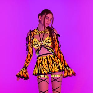 May include: A vibrant, eye-catching outfit featuring a crop top, skirt, and leg wraps. The ensemble showcases a butterfly-inspired design with orange and yellow patterns outlined in black. The long-sleeved top has flared sleeves, and the skirt is short. The leg wraps are black.
