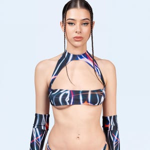 Op de afbeelding: Een zwart, futuristisch ensemble met een patroon van witte, oranje en blauwe lijnen. Het bestaat uit een haltertop, een bikinibroekje en armmouwen. Het model draagt de outfit tegen een lichtblauwe achtergrond.