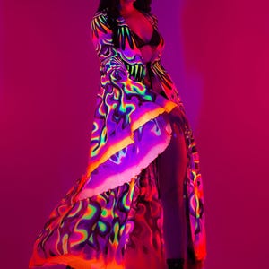 Könnte beinhalten: Ein langes, fließendes Gewand mit einem lebendigen, psychedelischen Muster in Neonfarben von Lila, Grün, Gelb und Orange. Das Gewand hat einen hohen Schlitz und wird mit schwarzen Stiefeln kombiniert. Der Hintergrund ist ein Farbverlauf aus Rosa und Lila.