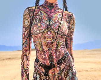 Könnte beinhalten: Ganzkörper-Jumpsuit mit langen Ärmeln und hohem Ausschnitt, mit einem lebendigen, psychedelischen Muster in Pink-, Lila-, Gelb- und Blautönen. Das Outfit umfasst einen schwarzen Gürtel mit Metallakzenten und wird mit einer schwarzen Sonnenbrille getragen. Der Hintergrund ist eine Wüstenlandschaft.