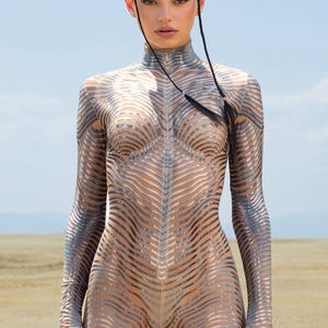 Puede incluir: Mono de color nude, de manga larga, con cuello alto y un patrón plateado en forma de ondas. El mono es ajustado y cubre todo el cuerpo. El fondo es un paisaje desértico bajo un cielo azul.