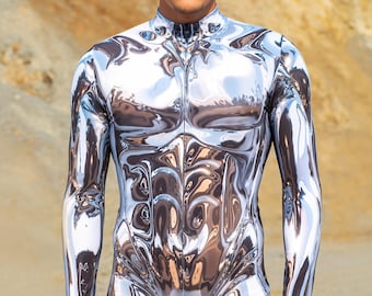 May include: A full-body silver metallic jumpsuit with a zipper closure. The long-sleeved garment has a reflective, liquid-like appearance, creating a sculpted effect. The model is standing in front of a sandy background.
