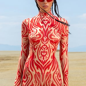 May include: A full-body jumpsuit with a high neckline and long sleeves. The jumpsuit features a red and beige tribal pattern. The model is wearing red sunglasses. The background is a desert landscape with a blue sky.
