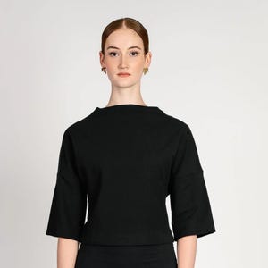 May include: A black top with a high neckline and three-quarter sleeves. The top is made of a solid, dark fabric and is worn by a person. The background is a plain white.