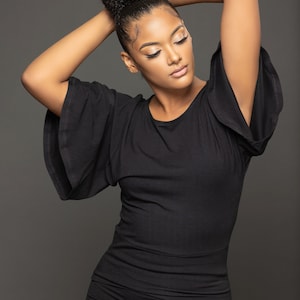 May include: A black dress with flared sleeves. The dress is form-fitting and reaches the knees. The model has her hair up in a bun and is posing with her arms above her head. The background is a solid gray.