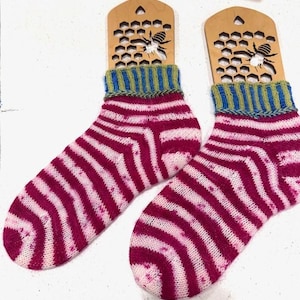 Könnte beinhalten: Ein Paar handgestrickte Socken mit einem gestreiften Muster in Rosa- und Weißtönen. Die Bündchen sind blau und grün. Jede Socke wird auf einem Sockenblocker aus Holz mit einem Bienen- und Wabenmuster präsentiert.