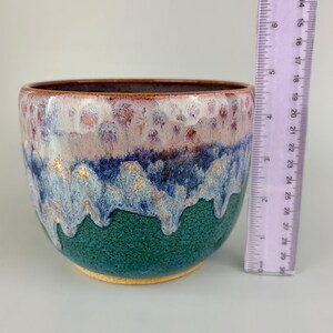 May include: A handmade ceramic bowl with a textured, glazed surface. The bowl features a color gradient, transitioning from a deep green base to blue, lavender, and brown at the rim. A ruler is placed next to the bowl for scale.