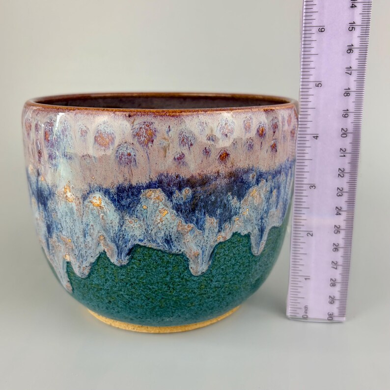 May include: A handmade ceramic bowl with a textured, glazed surface. The bowl features a color gradient, transitioning from a deep green base to blue, lavender, and brown at the rim. A ruler is placed next to the bowl for scale.
