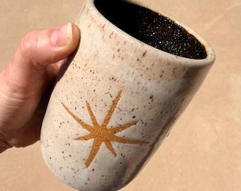 May include: A speckled ceramic mug with a starburst design in shades of tan and brown. The interior of the mug is dark brown. The mug is held at an angle, showcasing its cylindrical shape and the textured surface.