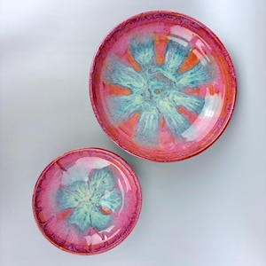 Può includere: Due ciotole in ceramica con un design vibrante e colorato. Le ciotole presentano un bordo rosa e arancione, con un centro turchese e giallo. La glassa crea una finitura lucida. Le ciotole sono di diverse dimensioni.