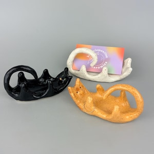May include: Three ceramic cat-shaped holders in black, orange, and white with speckles. The white holder displays a business card with the text 'koclementine ceramics'. Each holder features a curved tail and raised paws to support small items.