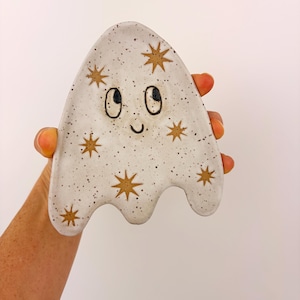 May include: A ghost-shaped ceramic trinket dish with a speckled white glaze. The dish features a hand-painted black face with two eyes and a small smile, accented by several gold-colored star motifs scattered across the surface.