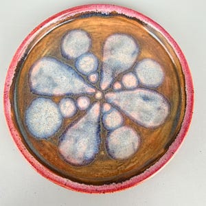 May include: A round ceramic plate featuring a brown glazed surface with a central floral pattern composed of light blue and white circular and teardrop-shaped crystalline glaze spots, finished with a vibrant pink rim around the outer edge.