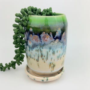 May include: A cylindrical ceramic planter with a matching saucer, featuring a reactive glaze that transitions from a bright green rim to deep blue, purple, and cream tones with vertical drip patterns and small gold-colored flecks throughout.