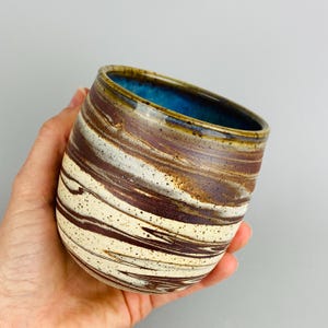 May include: A handmade ceramic tumbler with a unique marbled design. The exterior features swirling patterns of brown, cream, and white, with a speckled texture. The interior is a vibrant blue. The rim has a brown glaze.