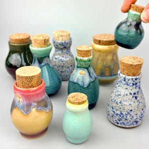 May include: A selection of small ceramic bottles with cork stoppers. The bottles are in various colours, including green, blue, yellow, and red, with speckled and glazed finishes. One bottle is being held by a hand.