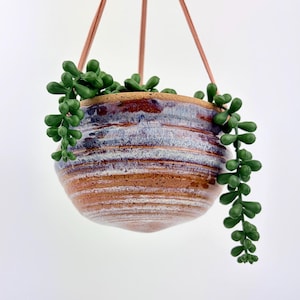 May include: A hanging ceramic planter with a rounded, ribbed shape featuring a glaze of blended brown, white, and deep purple tones. The planter is suspended by thin cords and contains a trailing green succulent plant with small, round leaves.