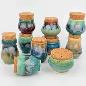 May include: A collection of eight small ceramic jars with cork lids, featuring various reactive glazes in shades of green, blue, yellow, and brown. Each jar has a rounded body and a smooth, glossy finish with unique color transitions and patterns.