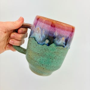 May include: A ceramic mug featuring a speckled light green base that transitions into a dark blue and vibrant magenta drip glaze at the rim. The mug has a sturdy handle and a smooth, glossy finish on the upper portion with a textured lower body.