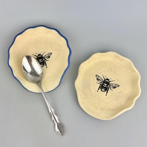 May include: Two round, cream-colored ceramic dishes with wavy edges, each featuring a black bee illustration in the center. One dish has a blue rim and holds a silver-toned metal spoon with a decorative handle resting inside the bowl.