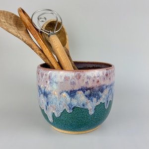May include: A ceramic utensil holder with a color gradient of green, blue, and purple. The holder contains wooden spoons and a whisk. The holder is a kitchen accessory.