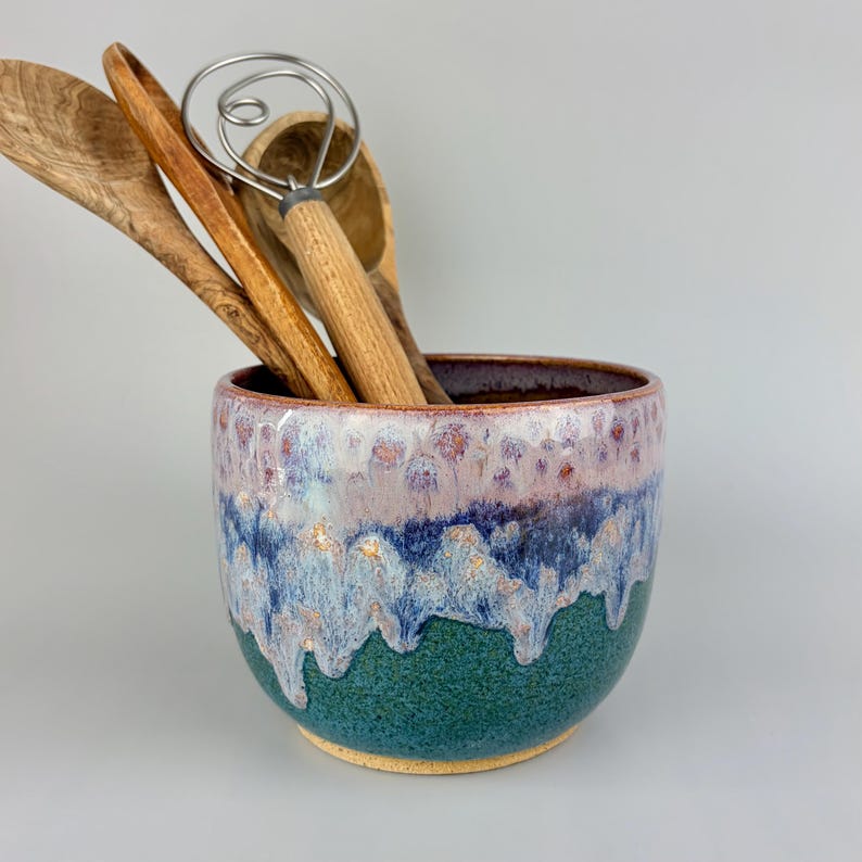 May include: A ceramic utensil holder with a color gradient of green, blue, and purple. The holder contains wooden spoons and a whisk. The holder is a kitchen accessory.