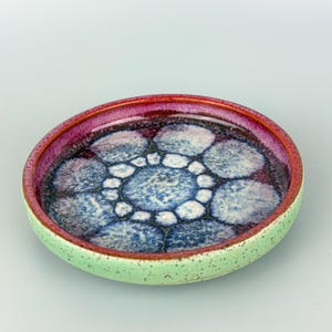 May include: A small, round ceramic dish with a speckled light green exterior. The rim is a deep red, and the interior features a pattern of blue and white circles. The dish is likely handmade and has a unique, artistic design.