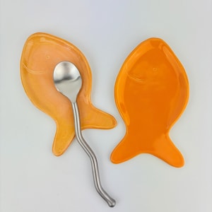 May include: Two fish-shaped ceramic spoon rests with a glossy finish. One spoon rest is a light orange, and the other is a vibrant orange. A silver-colored metal spoon with a long, wavy handle rests on top of the lighter orange fish-shaped dish.