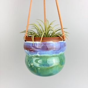 May include: A hanging ceramic planter with a gradient of colors, including green, blue, and purple. The planter is suspended by tan leather straps and holds a small air plant. The rim of the planter is brown.