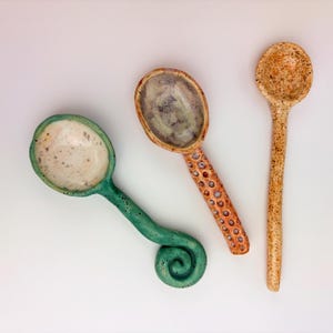 May include: Three handmade ceramic spoons with distinct designs. One features a green speckled finish and a spiral handle, another has a textured orange honeycomb handle with a purple bowl, and the third is a long, slender, tan speckled spoon.