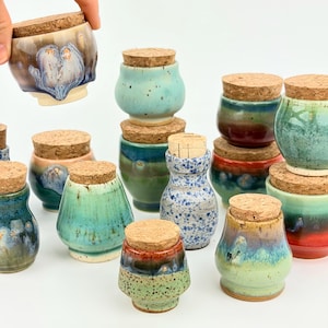 May include: A collection of small ceramic jars with cork stoppers, featuring various glaze colors including shades of blue, green, red, and brown. The jars have rounded or tapered shapes with smooth, glossy, or speckled finishes and textured surfaces.