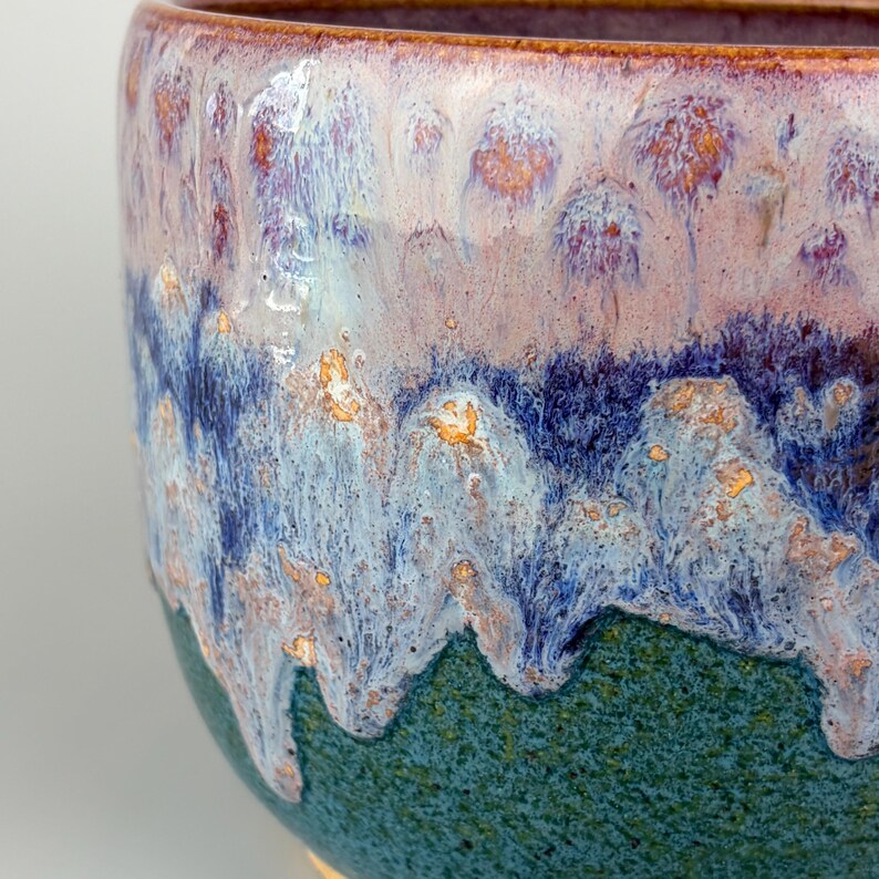 May include: A handcrafted ceramic bowl with a unique glaze. The bowl features a gradient of colors, including teal, blue, purple, and white, with gold accents. The interior rim is a warm brown. This decorative bowl is suitable for serving or display.