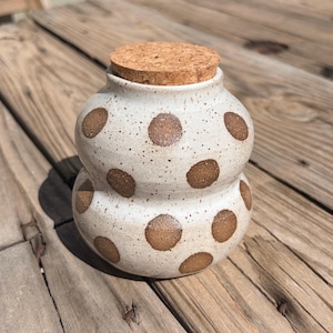 May include: A speckled ceramic jar with a cork lid. The jar is off-white with large brown polka dots. The jar has a bulbous shape with a smaller base and a wider top. The cork lid is round and fits snugly on the jar.