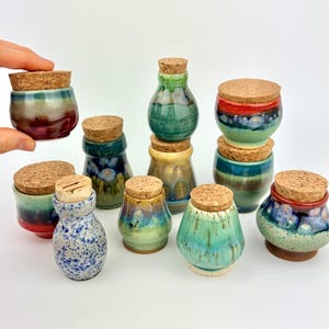 May include: A collection of small, hand-thrown ceramic jars with cork stoppers, featuring various glaze colors including shades of green, blue, red, and speckled white. Each jar has a unique shape and finish, displayed together on a plain surface.