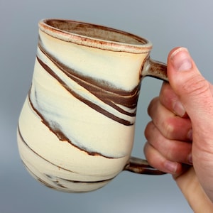 May include: Cream-colored ceramic mug with brown marbling and a brown handle. The mug has a unique shape and is held by a hand. The mug is a handmade item and is perfect for coffee or tea.