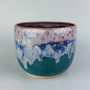 May include: A handcrafted ceramic bowl with a unique glaze. The bowl features a dark teal base, transitioning to a blue and white middle section with a dripped effect, and a purple and brown rim. The interior is a dark brown color.