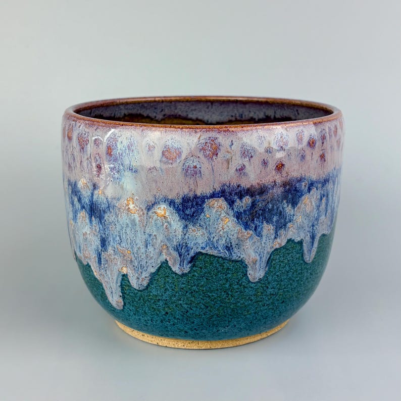 May include: A handcrafted ceramic bowl with a unique glaze. The bowl features a dark teal base, transitioning to a blue and white middle section with a dripped effect, and a purple and brown rim. The interior is a dark brown color.