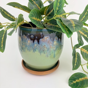 May include: A round ceramic planter with a glossy glaze that transitions from a light sage green base to a dark blue and brown drip pattern at the rim. The planter sits on a matching terracotta-colored saucer and contains a green leafy plant.