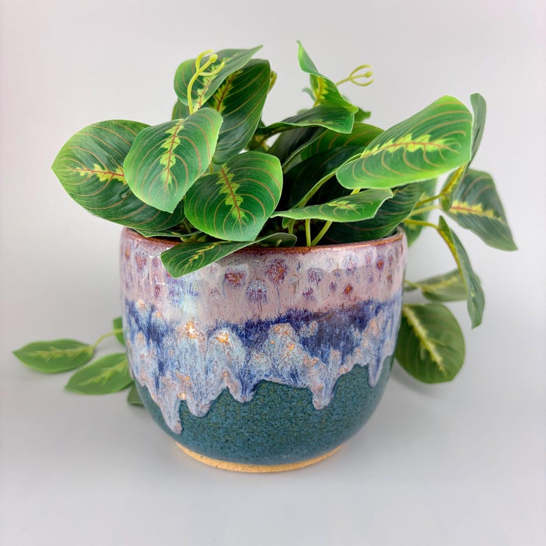 May include: A ceramic plant pot with a green, blue, and purple glaze. The pot is filled with a green plant with large, veined leaves. The pot has a round shape and a textured surface.
