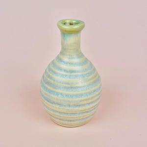May include: A ceramic vase with a rounded body and a narrow neck. The vase features horizontal bands of light blue and cream colors. The rim of the vase is a pale green. The vase is set against a pink background.