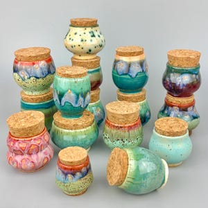 May include: A collection of small ceramic jars with cork stoppers, featuring various glaze finishes including speckled cream, vibrant turquoise, deep blue, and pink. Each jar has a rounded body and a fitted natural cork lid for closure.