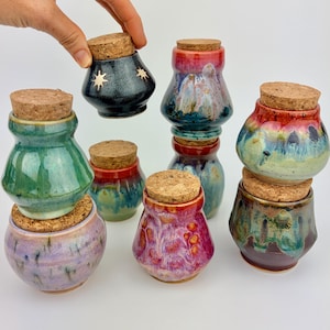 May include: A collection of small, bulbous ceramic jars with cork stoppers, featuring various glossy, multi-colored glazes including green, purple, red, and blue. One dark jar is decorated with gold star motifs, while others show blended, drip-style glaze patterns.