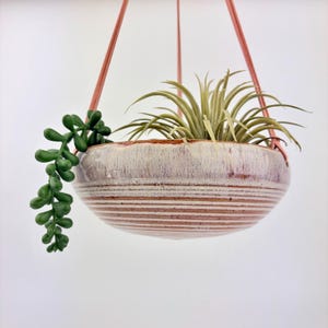 May include: A hanging ceramic planter with a rounded bowl shape, featuring a white and reddish-brown glazed finish with horizontal ridges. The planter is suspended by thin pink cords and contains a spiky air plant and a trailing green succulent.