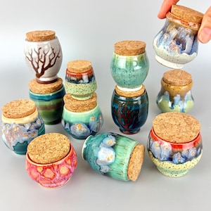 May include: A collection of small, handcrafted ceramic jars with cork lids. The jars feature a variety of colors and designs, including blue, green, red, and white, with unique patterns and textures. The jars are of varying shapes and sizes.