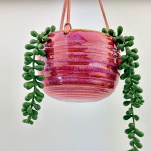 May include: A round, ribbed ceramic hanging planter with a glossy, variegated magenta and pink glaze. The planter is suspended by thin, light-colored cords and contains a trailing artificial green succulent plant with small, bead-like leaves.