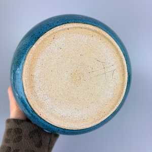 May include: A handmade ceramic bowl with a teal blue glazed exterior and a natural, unglazed interior. The bowl's base is visible, showing the raw, textured clay. The bowl is held by a person.