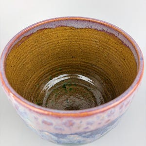 May include: A handmade ceramic bowl with a textured interior and a glossy, multi-colored exterior. The bowl features a brown interior, a purple rim, and a blue and purple exterior with a textured pattern. The bowl is a unique piece of pottery.