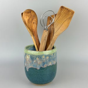 May include: A ceramic utensil holder with a blue and green drip glaze finish containing a wooden spoon, a wooden spatula, a wooden flat turner, and a metal wire whisk with a wooden handle. The container has a cylindrical shape.